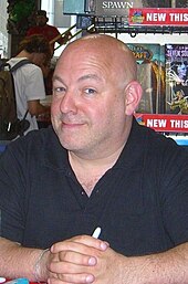 Brian Michael Bendis seated, smiling with his hands folded at a comic book convention