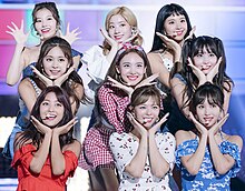 An image of Twice doing the same pose with their hands except for Sana.