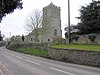 Stone building with square tower, separated from the road in the foreground by a stone wall.