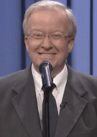 Headshot of an elderly man, wearing glasses and a morning suit, speaking at an event.