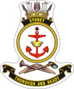 A ship's badge. A naval crown sits on top of a black scroll with "SYDNEY" written in gold. This is atop a yellow, rope-patterned ring, in which a red anchor is centred. Below the ring are a stone axe and a nulla nulla sitting on top of a boomerang. At the bottom of the badge is a black scroll with "THOROUGH AND READY" written.