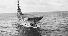 An aircraft carrier underway at sea. The bow of the aircraft carrier has been torn off, and the surrounding area is damaged and scorched.