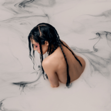 A naked woman is shown from behind, submerged in a pool of white liquid with black wisps.