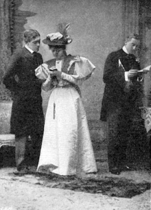 young white male and female couple in Victorian costume conversing; she writes in a small notebook, left, while a young white male, right, listens to them, secretly, and writes in his notebook