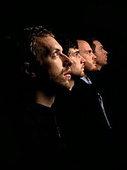 Coldplay facing to the right in profile position with a dark background behind them